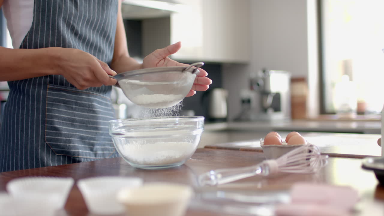 Happy biracial woman biracial woman in apron sieving flour, baking in kitchen, slow motion