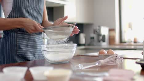 happy biracial woman biracial woman in apron sieving flour, baking in kitchen, slow motion