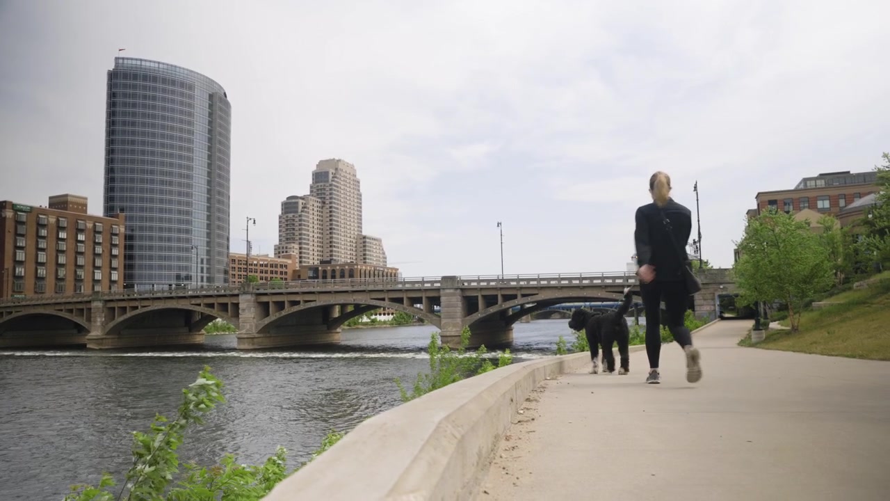 Ângulo baixo, filmagem feita à mão de uma jovem passeando com seu cachorro ao longo de um caminho de rio em um ambiente urbano. Filmado em 4k