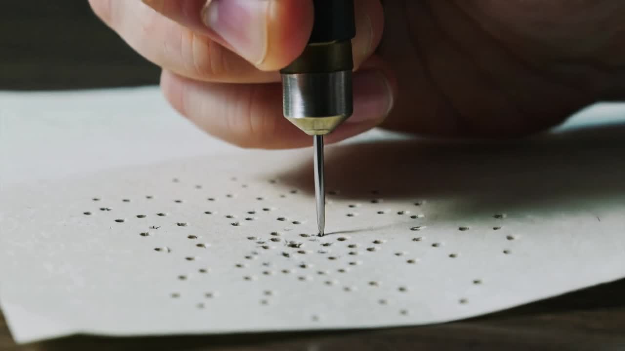 Mano adulta che si abbassa, usando un punteruolo per forare della carta bianca su un tavolo di legno, creando un motivo a spirale di fori.