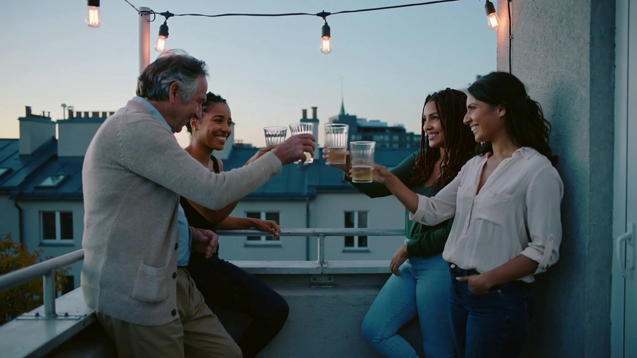 Lifting cup, four friends toasting clear cups on balcony at dusk under string lights, celebrating
