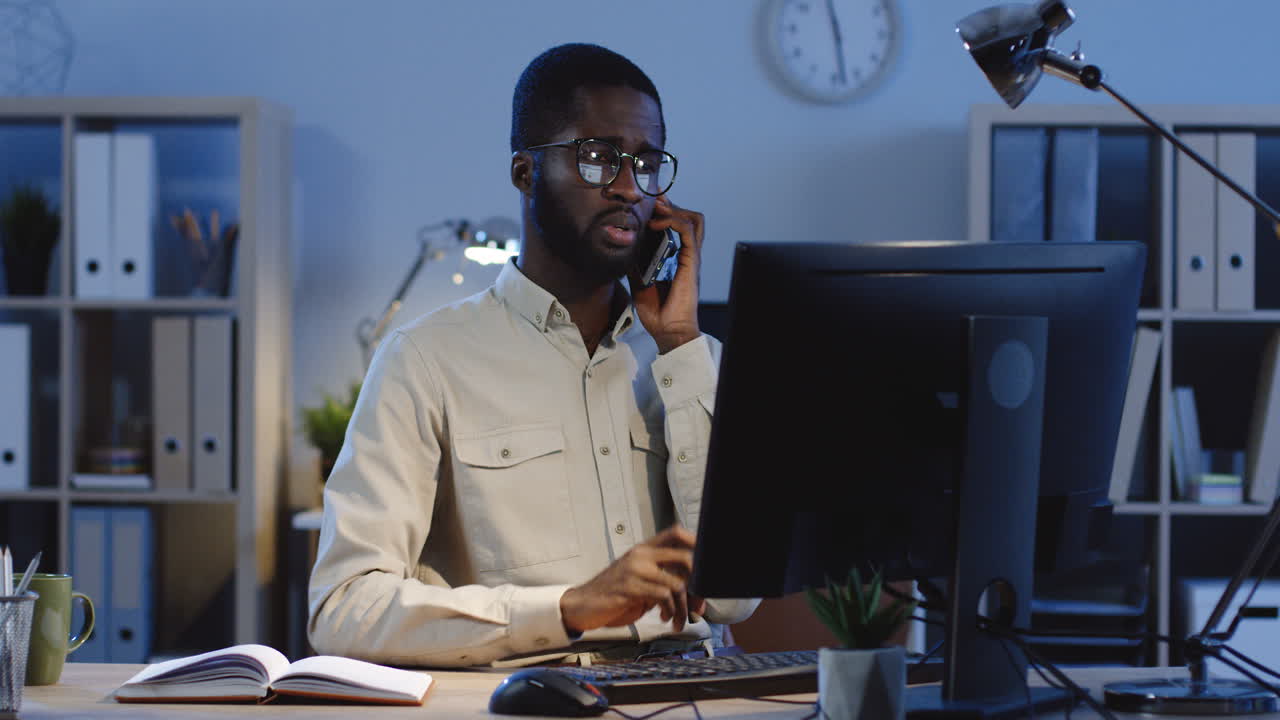 trabajador de oficina serio hablando por teléfono sentado frente al monitor de la computadora en la oficina por la noche