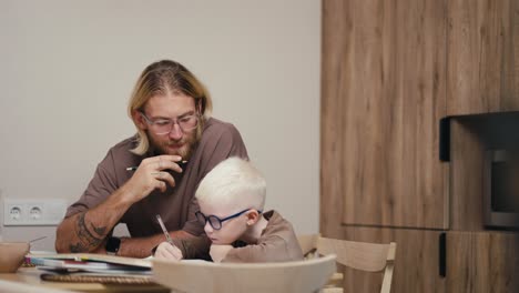 Ein-Blonder-Mann-Mit-Bart-Und-Brille-Hilft-Seinem-Kleinen-Albino-Sohn-Mit-Weißer-Haarfarbe-Und-Blauer-Brille,-Seine-Hausaufgaben-Zu-Machen-Und-Alles-Richtig-In-Mathematik-Zu-Berechnen,-Während-Er-Zu-Hause-In-Einer-Modernen-Küche-Hausaufgaben-Macht