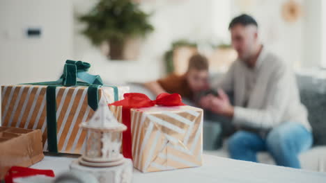 close up on christmas presents on table with dad and little boy in background playing