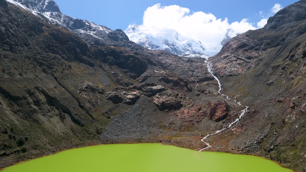 An aerial shot starts over the vibrant green water of Laguna Shallap, then tilts up to reveal a winding mountain stream flowing from snow-capped glaciers in the Cordillera Blanca, Peru