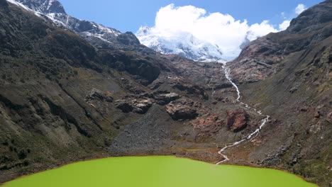 An aerial shot starts over the vibrant green water of Laguna Shallap, then tilts up to reveal a winding mountain stream flowing from snow-capped glaciers in the Cordillera Blanca, Peru