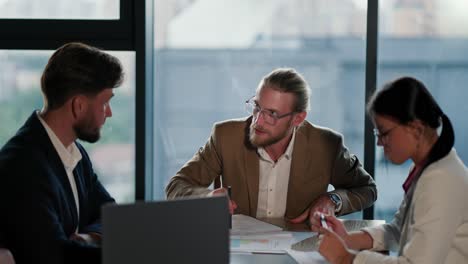 An-angry-office-worker,-a-blond-man-with-glasses-with-a-beard-in-a-light-brown-jacket,-hits-the-table-with-a-ballpoint-pen-and-shouts-at-his-employees.-Conversation-in-a-raised-voice-while-working-in-a-modern-office-with-a-panoramic-window