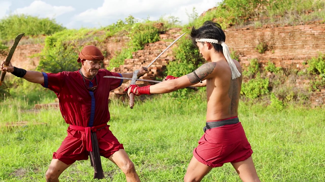 Dois homens com trajes tradicionais vermelhos praticam artes marciais com espadas perto de ruínas históricas de tijolos.