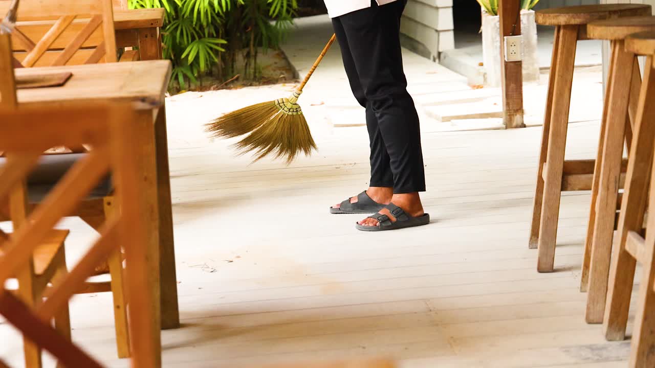 A person sweeps a wooden deck with a broom, surrounded by wooden furniture and greenery, under natural daylight