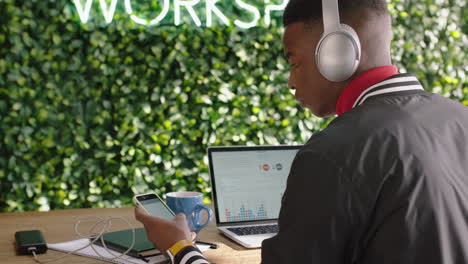 Joven-Afroamericano-Estudiante-De-Negocios-Usando-Un-Teléfono-Inteligente-Navegando-Por-Las-Redes-Sociales-Enviando-Mensajes-De-Texto-Disfrutando-De-Un-Estudio-En-Línea-Escuchando-Música-Usando-Auriculares