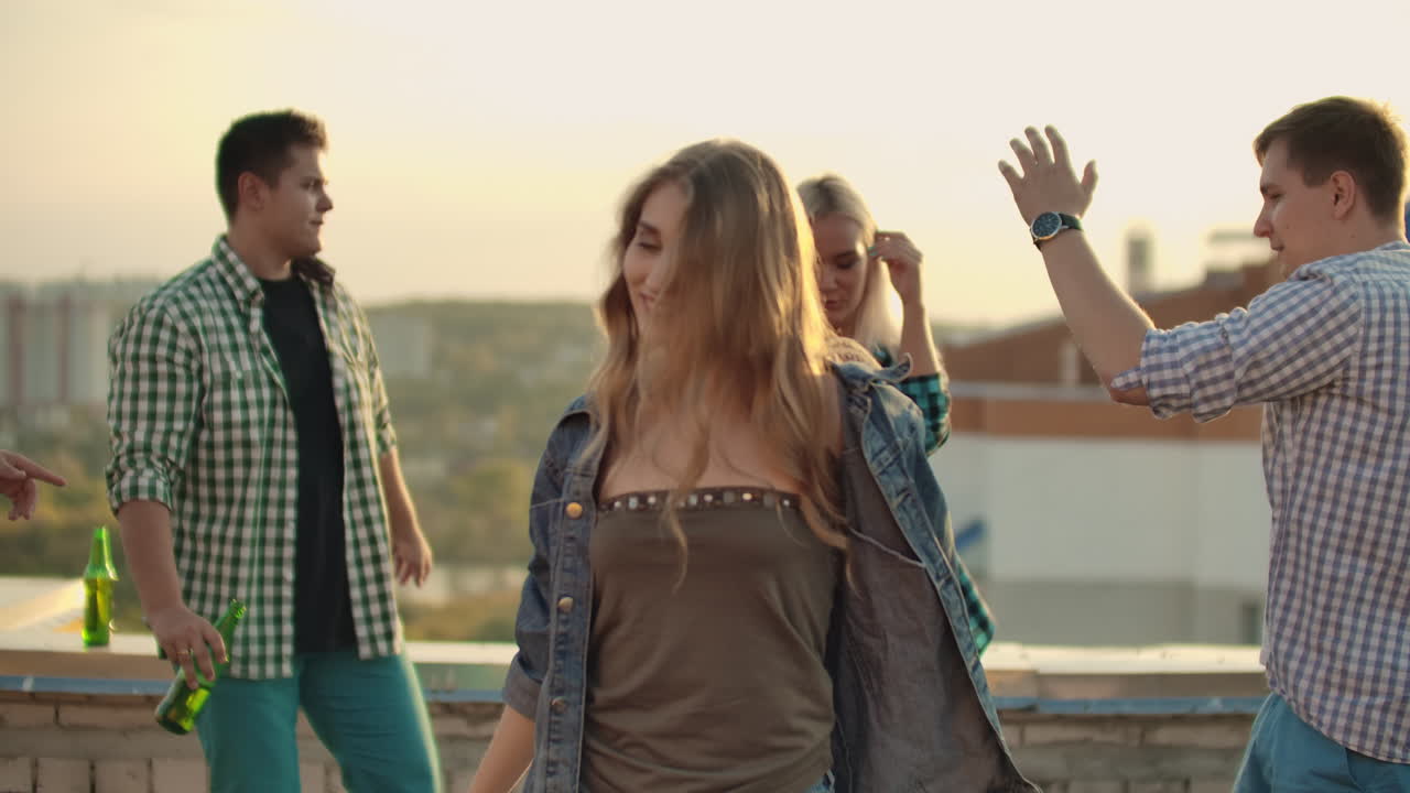 la joven y hermosa chica estadounidense está bailando en el techo con sus cuatro amigas en la fiesta. ella sonríe y disfruta del tiempo en pantalones cortos y una chaqueta de denim ligera en la noche de verano. su cabello rubio está volando en el viento.