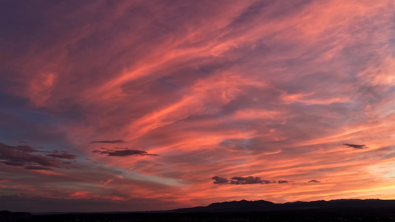 Timelapse 4K UHD con dron: vibrantes colores rosa y naranja del atardecer iluminan amplias formaciones de nubes plumosas. Un impresionante cielo crepuscular desértico transiciona sobre una cordillera silueteada.