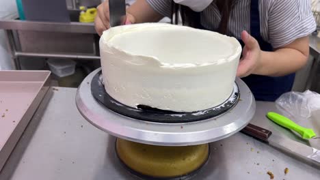 chef de repostería femenina usando un raspador de torta alisando el glaseado de merengue de crema de mantequilla en el lado de la torta, y girando lentamente el puesto de torta en un entorno de panadería comercial