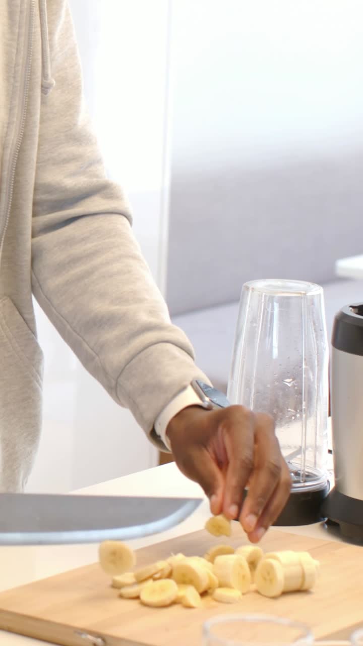 Video verticale: Uomo incappucciato che guarda in basso, taglia una banana per frullarla in cucina, bloccando la tazza; con spazio per il testo.