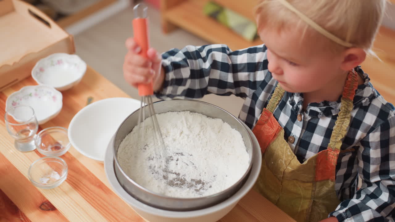 Young child in apron uses whisk to sieve flour into bowl on wooden table while another child partially visible nearby, surrounded by apples, bowls, and utensils during kitchen cooking activity