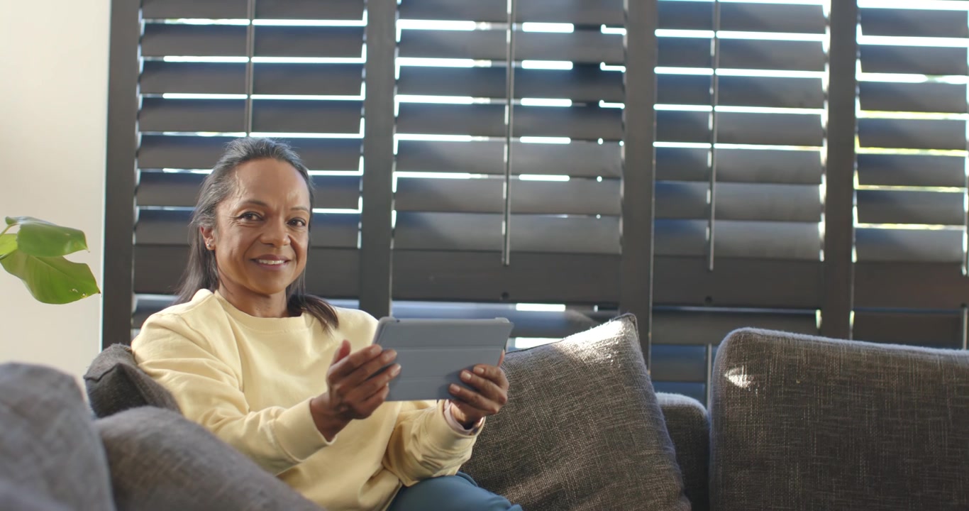 African American woman sitting on sofa at home viewing tablet, shifting and smiling