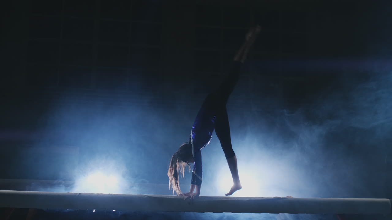 la niña es una atleta profesional realiza un truco acrobático de gimnasia en una viga con luz de fondo y cámara lenta en ropa de gimnasia deportiva. humo y azul. saltar y girar en la viga de equilibrio