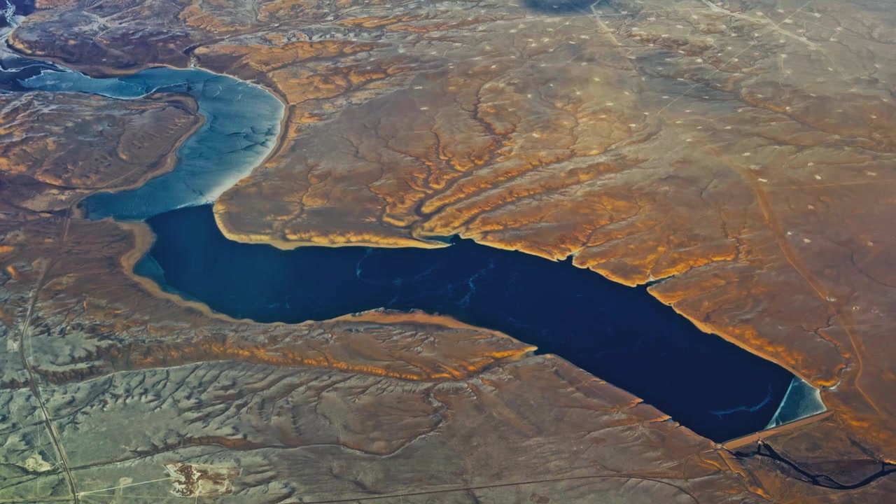 Aerial of winding reservoir and arid terrain seen from airplane window, lush green orange pockets along edge of dam reservoir