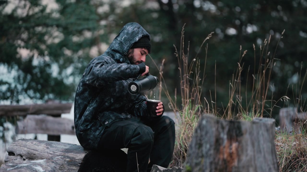 En mand i en hættetrøje hælder varmt vand i en mate-kop, mens han sidder på en træstamme i en skov. Ideel til brug i udendørs livsstils- eller campingrelateret indhold.