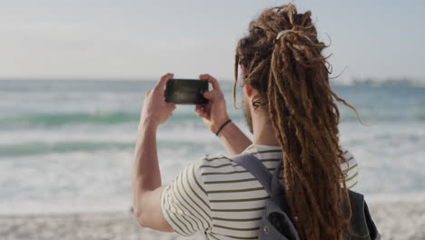 young tourist man taking photo on beach using smartphone camera technology of beautiful scenic ocean background caucasian male enjoying summer vacation travel