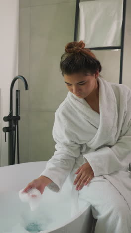 una mujer toca la espuma sentada en el borde de la bañera. una joven atractiva en vestido de terry juega con espuma suave en un baño elegante. procedimiento de salón de spa