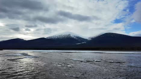 Schneebedeckte-Berge-Unter-Einem-Bewölkten-Himmel,-Mit-Einem-Teilweise-Gefrorenen-Fluss-Im-Vordergrund