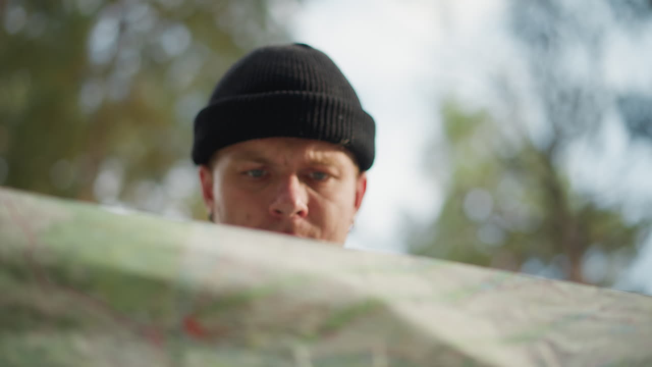 White hiker studies map in forest, wearing black beanie and backpack while scanning horizon for route acts as navigator and trail scout, checks compass, senses sunlight through pines, focused