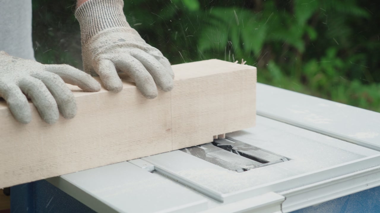 Close-up of a carpenter's hands in gloves guiding a large wooden beam through a table saw. Sawdust flies as the blade cuts the timber outdoors.