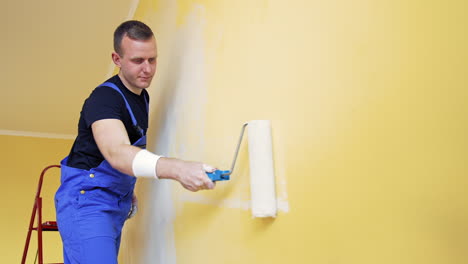 room makeover. portrait of a young man who painting wall. worker painting wall in new color with a paint roller from a ladder indoors.