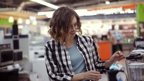 in the appliances store, a brunette curly woman in a plaid shirt chooses a blender for shopping by viewing and holding the