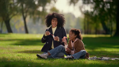 Happy-daughter-blowing-soap-bubble-sitting-in-sunny-park.-Joyful-family-weekend.