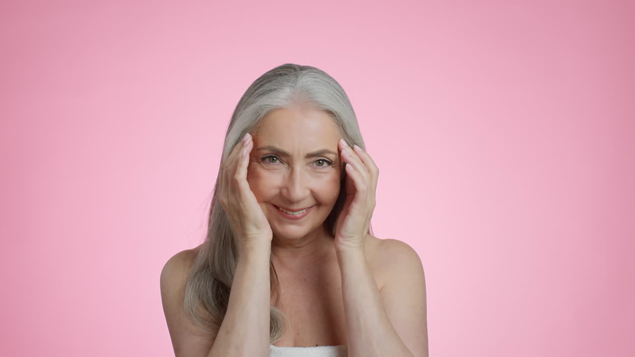 Smiling Senior Woman Demonstrating Skincare on Pink Background
