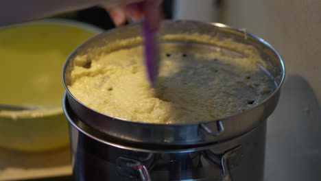 hand uses plastic spatula in pressing potato dough batter onto spaetzle maker over pot with boiling water. close up