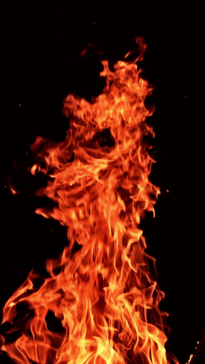 Huge burning fire with tiny sparkles flying up quickly. Flame of big bonfire striving up at black backdrop. Vertical video