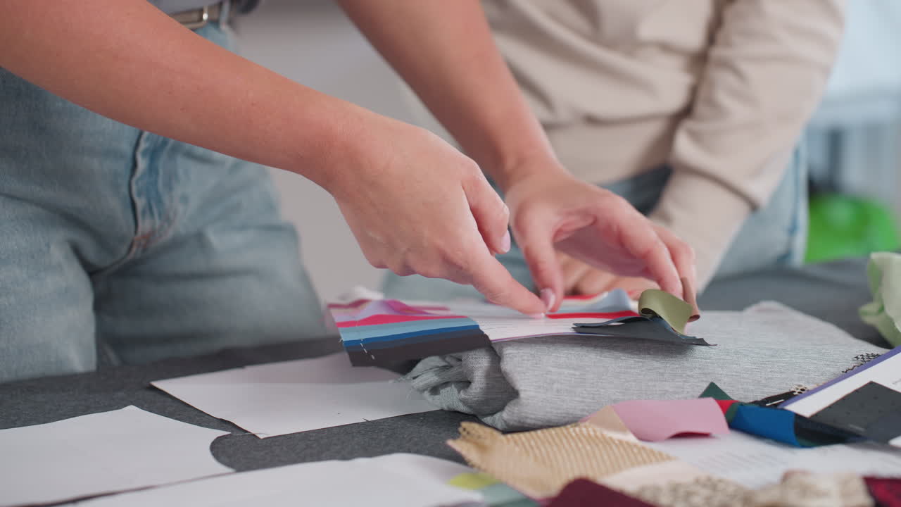 Female stylists comparing colorful fabric swatches on work table filled with vibrant materials including blue red yellow green samples during creative session in bright fashion studio