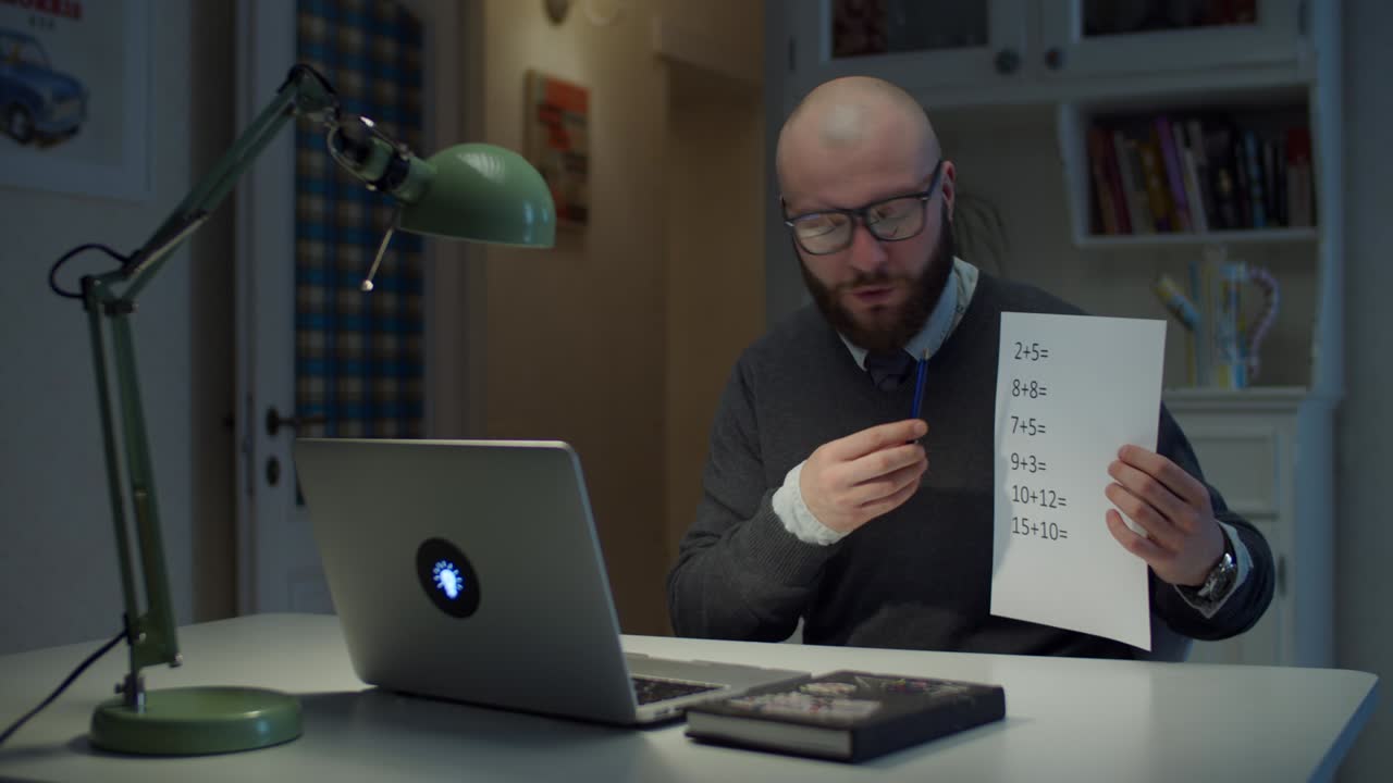hombre barbudo de 30 años con gafas mostrando una hoja de papel con ecuaciones matemáticas simples mirando una computadora portátil en casa. proceso de educación en línea. vista lateral del maestro hablando.