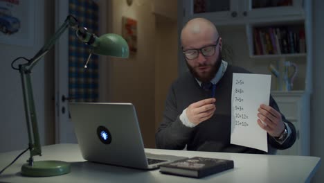 hombre barbudo de 30 años con gafas mostrando una hoja de papel con ecuaciones matemáticas simples mirando una computadora portátil en casa. proceso de educación en línea. vista lateral del maestro hablando.