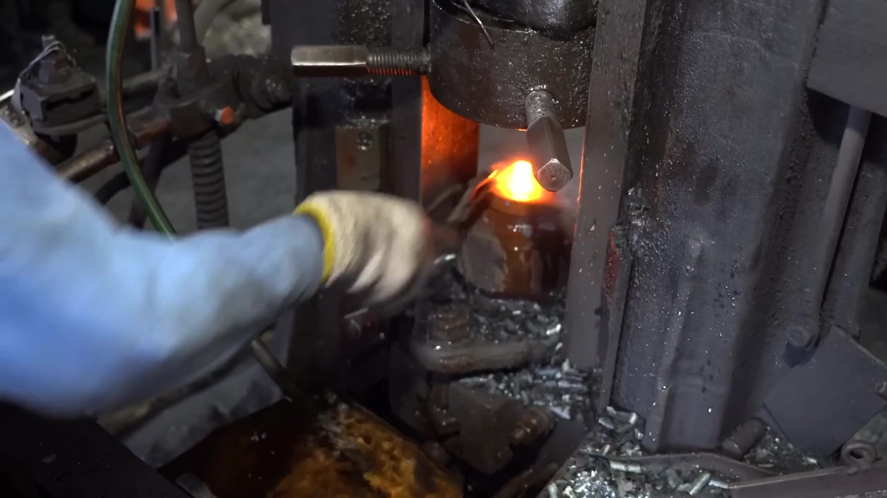 Worker using tongs to pull a glowing forged bolt head from the press die