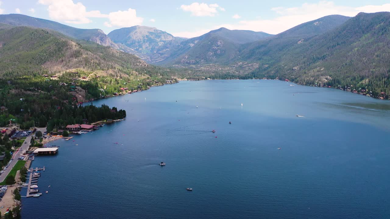 Panorama aereo di un lago alpino con porticcioli, sviluppo costiero, pendii boscosi e cime montuose distanti riflesse in ampie acque calme in una soleggiata giornata estiva.