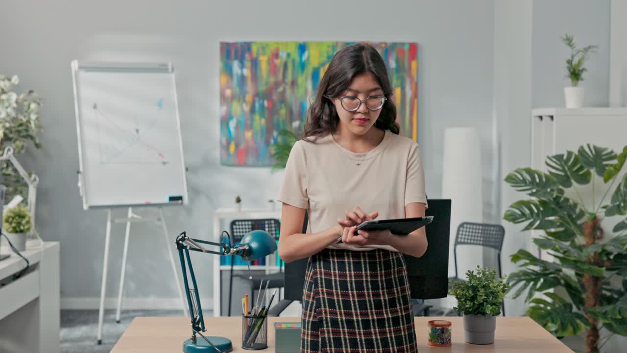 una hermosa mujer con gafas está de pie frente al escritorio en el medio de la oficina, comprobando en la tableta las órdenes de la compañía, el número de órdenes a completar, asignando tareas a los compañeros de trabajo