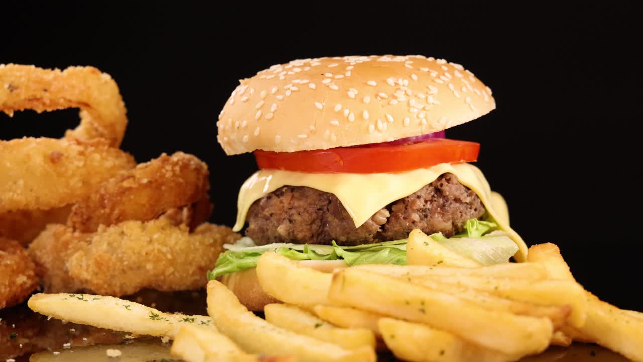 Juicy beef burger with lettuce, tomato, cheese, onion rings, and French fries rotates on a black background under bright studio lighting, highlighting textures