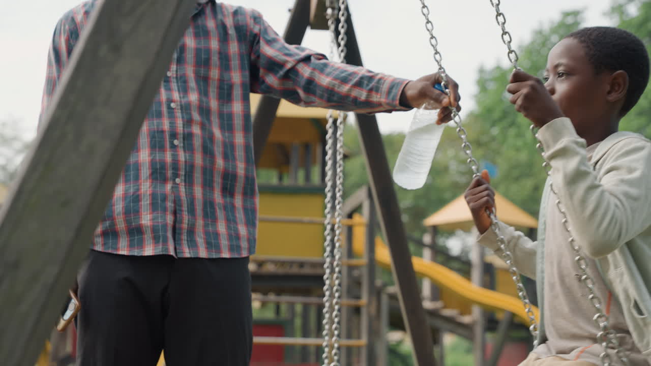 Ayah menawarkan air kepada putra di ayunan di taman, ayah berkotak-kotak memegang botol saat anak minum, rantai dan perosotan taman bermain di latar belakang, momen kehangatan dan ikatan yang tenang