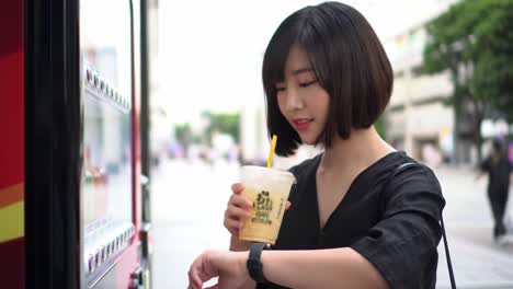 a young woman enjoys her refreshing drink while using a vending machine on a sunny day in an urban environment, capturing delight and anticipation.