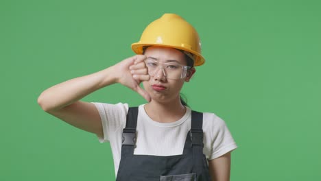 close up of asian woman worker wearing goggles and safety helmet showing thumbs down gesture to camera while standing in the green screen background studio