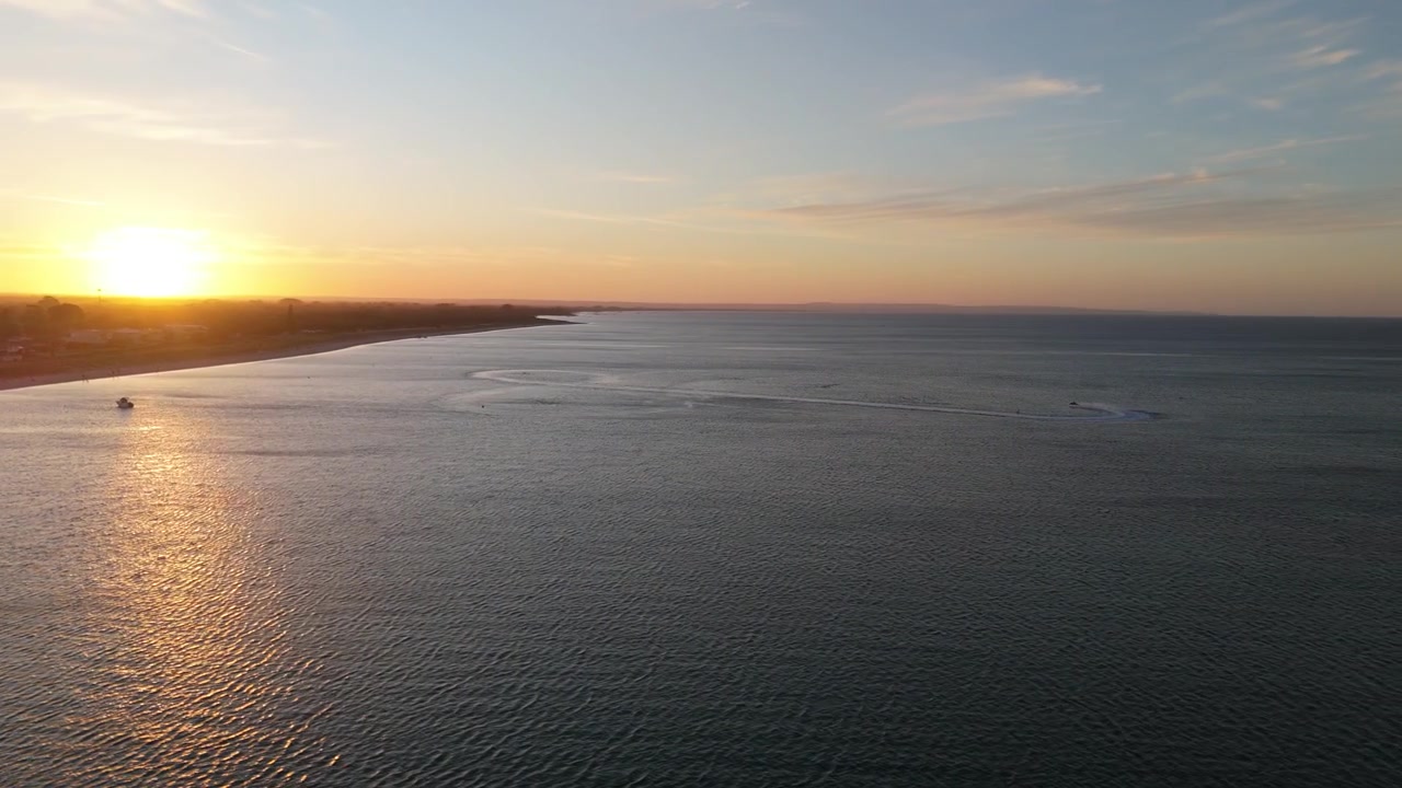 Un tramonto sereno che riflette una calda luce dorata sull'oceano con una moto d'acqua che si muove sull'acqua leggermente increspata, mentre una barca all'ancora riposa in lontananza
