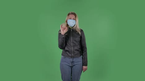 portrait of beautiful young woman in medical mask is looking straight sighs relieved and making sign ok. blonde with loose hair in a black jacket and jeans on a green screen in the studio. health protection corona virus concept