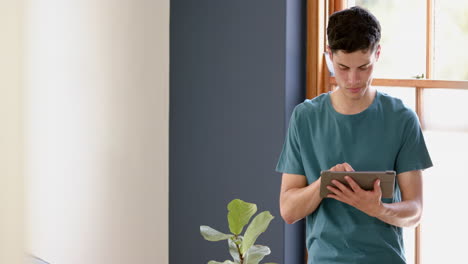 Focused-biracial-man-using-tablet-standing-in-sunny-living-room,-copy-space,-slow-motion