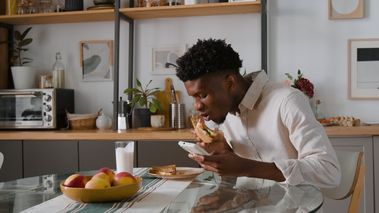 Man eating sandwich while using phone in the kitchen