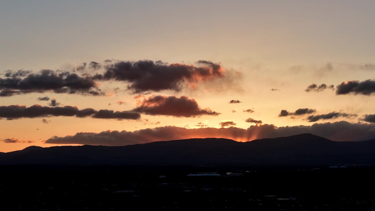 4K drone timelapse captures a rippling orange sunset glow behind a dark mountainside. Cinematic flying motion tracks fast moving clouds over a vast southwestern landscape under an evening afterglow.
