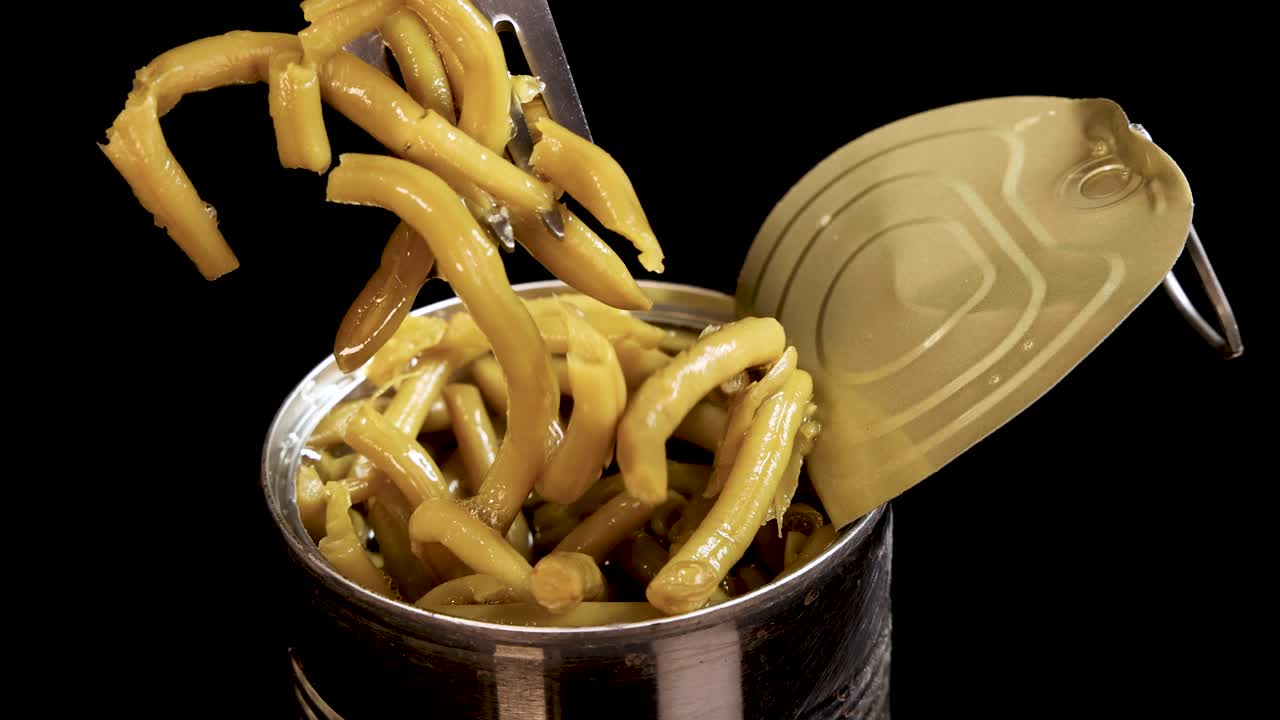 A metal fork lifts cooked haricot beans from an open tin can against a black background, with dramatic studio lighting and close-up camera angles